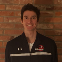 Man in a dark jacket with a logo stands against a brick wall