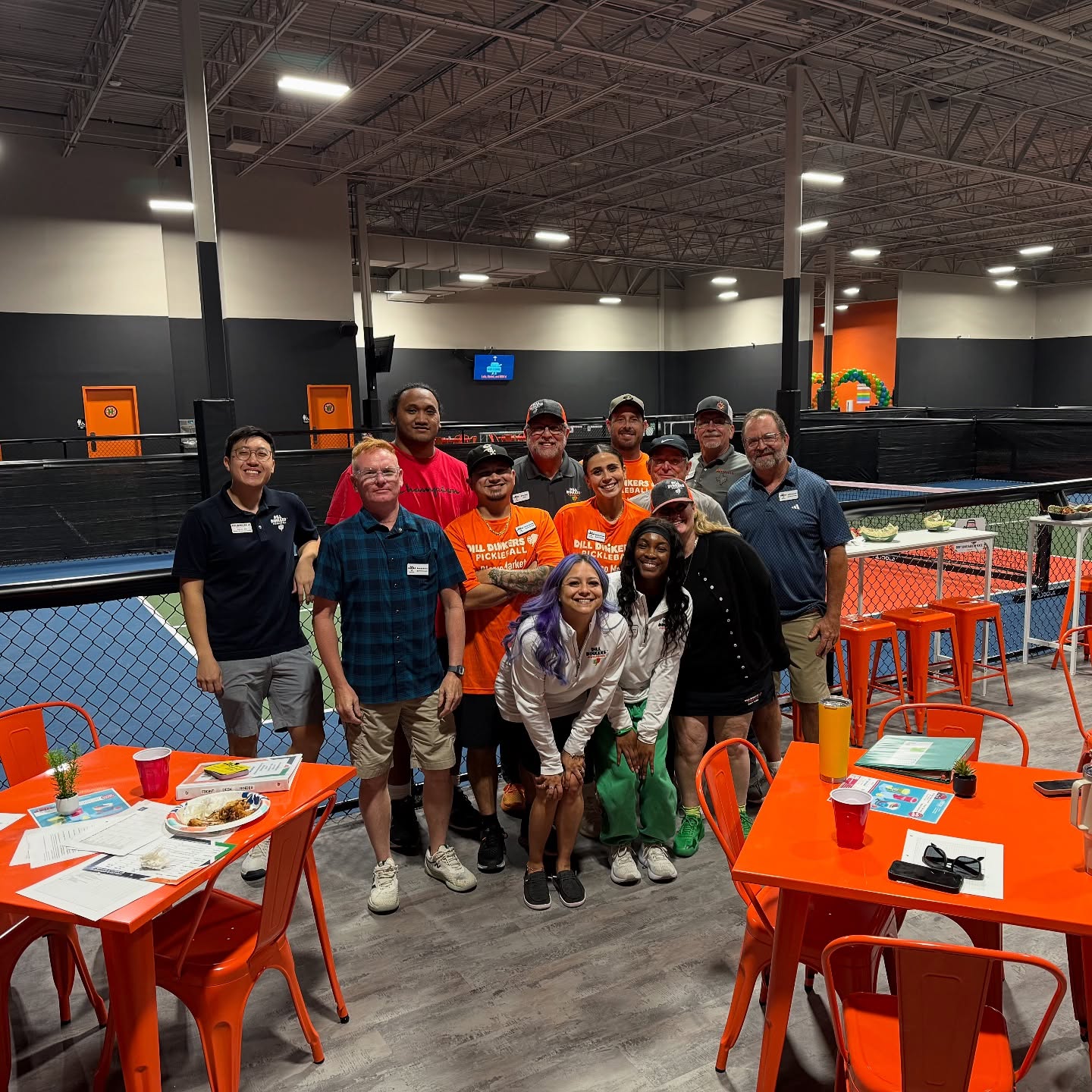 497901497_17879364819313701_648681768634991155_n Group of people posing on an indoor court, orange chairs and tables in foreground