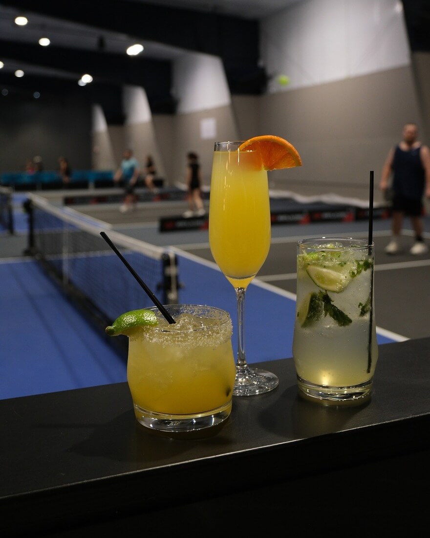 Three cocktails sit on the bar overlooking the pickleball courts
