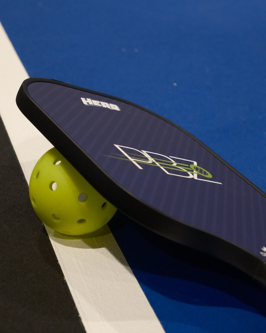 A Pickleball Zone branded pickleball paddle laid on a pickleball court with a pickleball ball.
