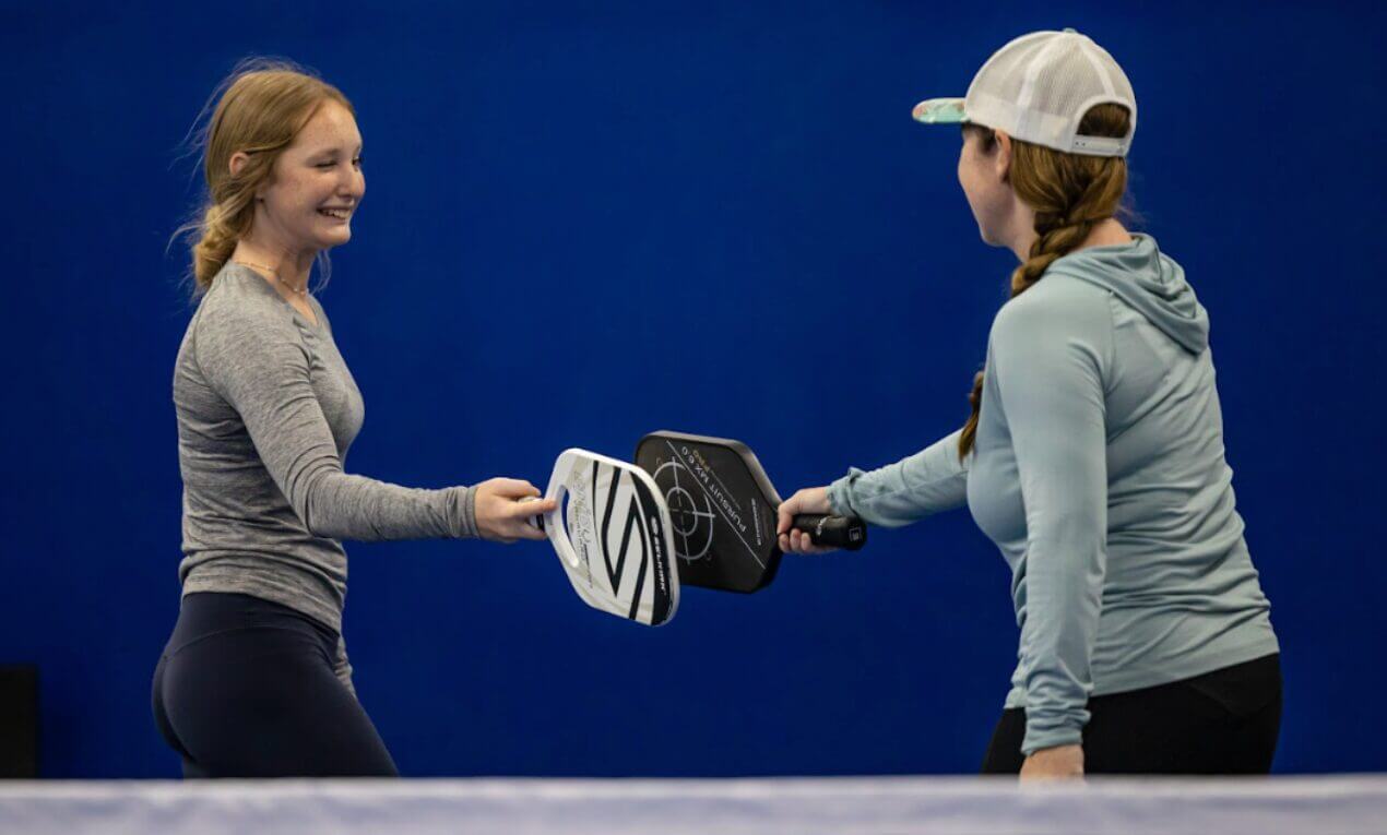 Two women on an indoor pickleball court tap paddles at the net in a friendly gesture after a game. One wears a gray long-sleeve shirt and the other wears a light blue hoodie and cap, both smiling with a bright blue wall in the background.