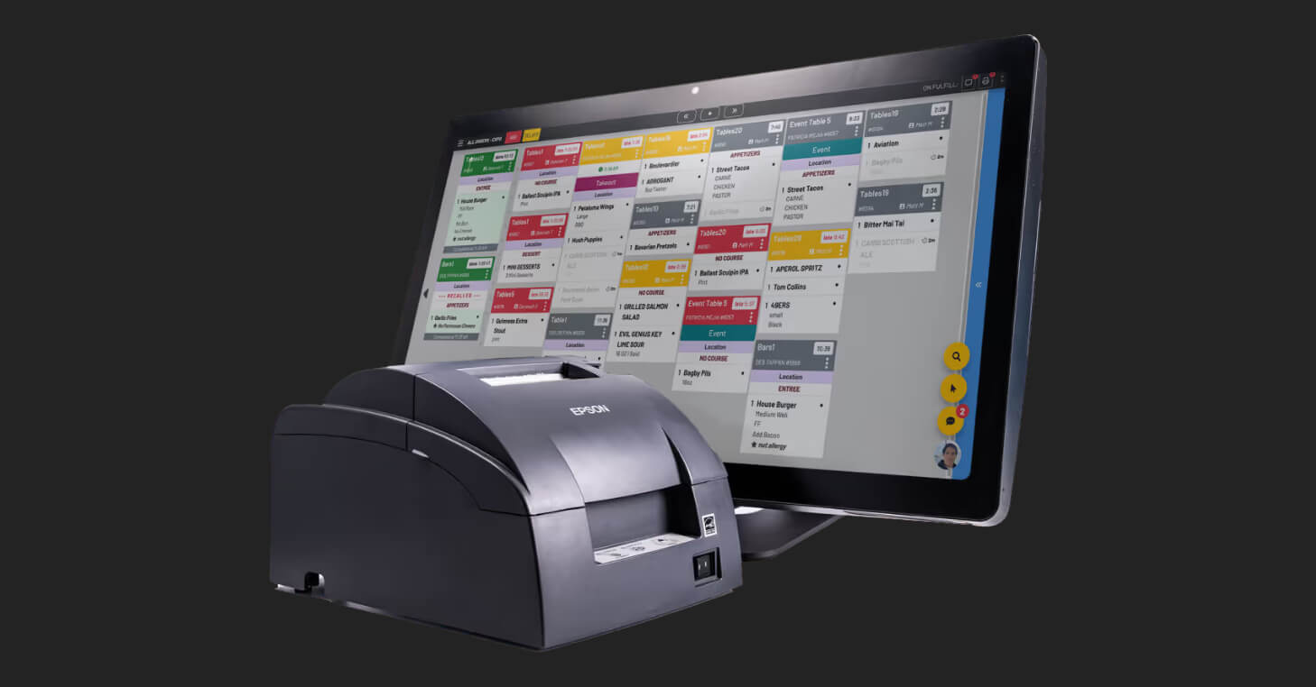 A kitchen display system featuring a touchscreen monitor and an Epson receipt printer. The screen shows a detailed order management interface with color-coded tickets for various tables and menu items, illustrating a digital solution for streamlining back-of-house restaurant operations.