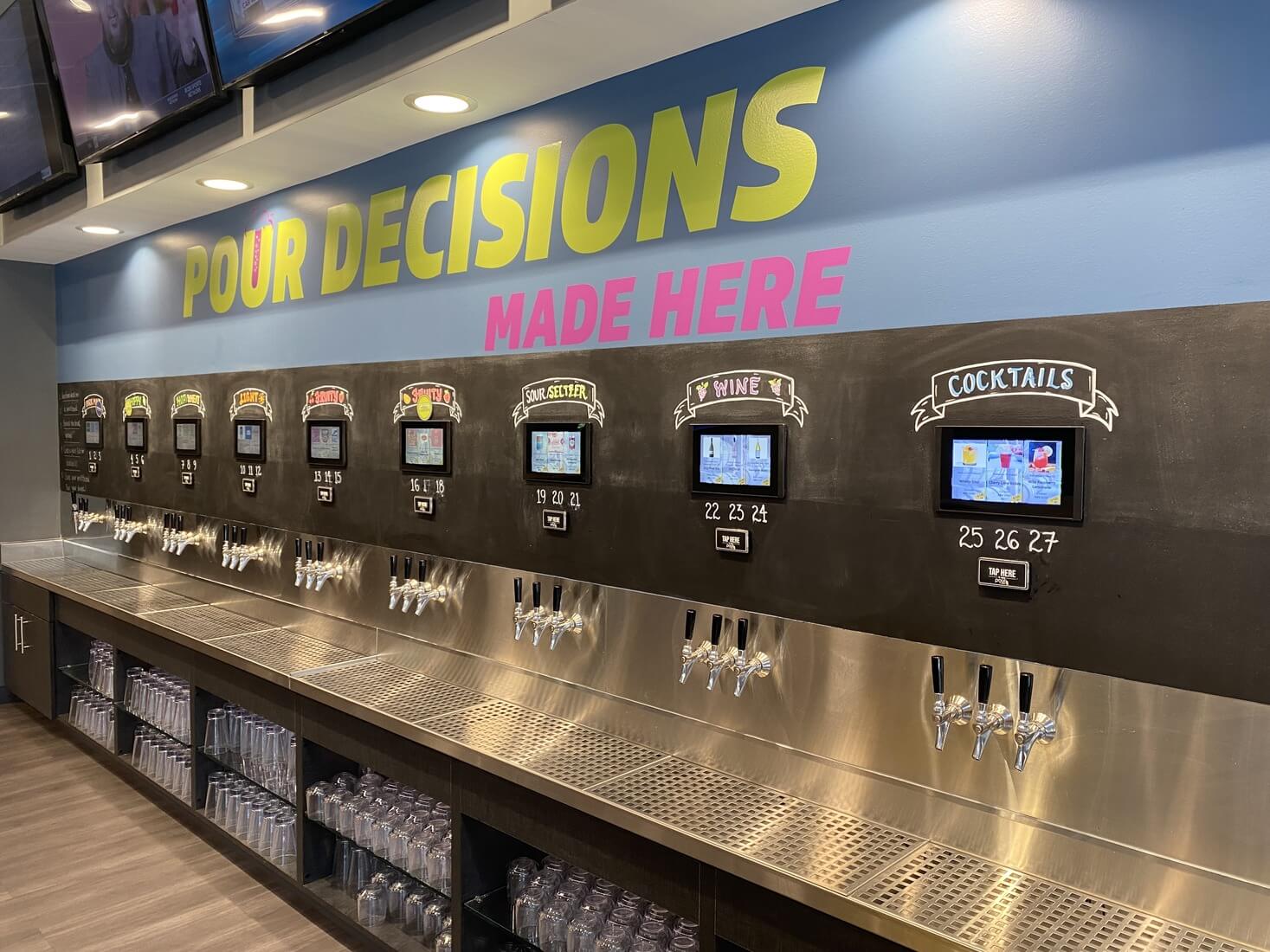 A modern self-serve beverage wall with multiple taps and digital screens labeled by drink type, including beer, cider, seltzer, wine, and cocktails. Above the stations, a bold sign reads “POUR DECISIONS MADE HERE” in colorful lettering. Dozens of clear plastic cups are neatly stacked below the taps, ready for use.
