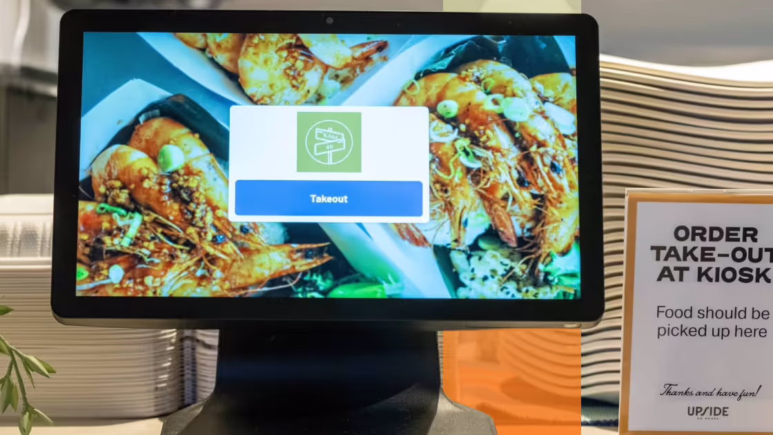 A touchscreen kiosk displays a vivid image of shrimp dishes with a “Takeout” button in the center. To the right, a sign instructs customers to “ORDER TAKE-OUT AT KIOSK” and notes that food will be picked up nearby. Plates are stacked in the background, reinforcing the restaurant's self-service system.