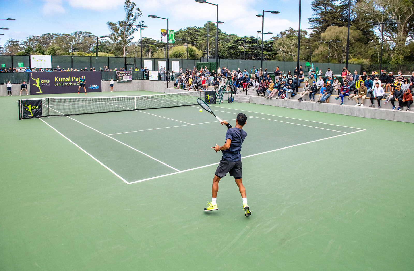 Tennis match at a municipal facility with spectators watching from stands.