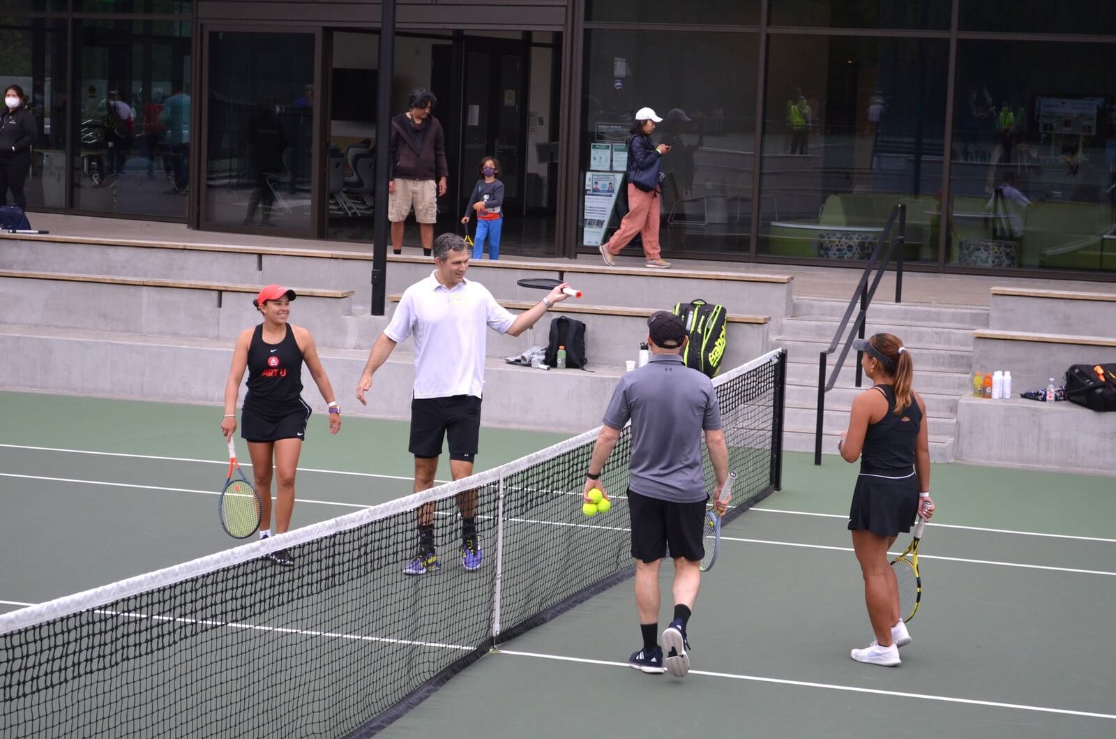 Group of tennis players gathered at the net in front of a modern clubhouse.