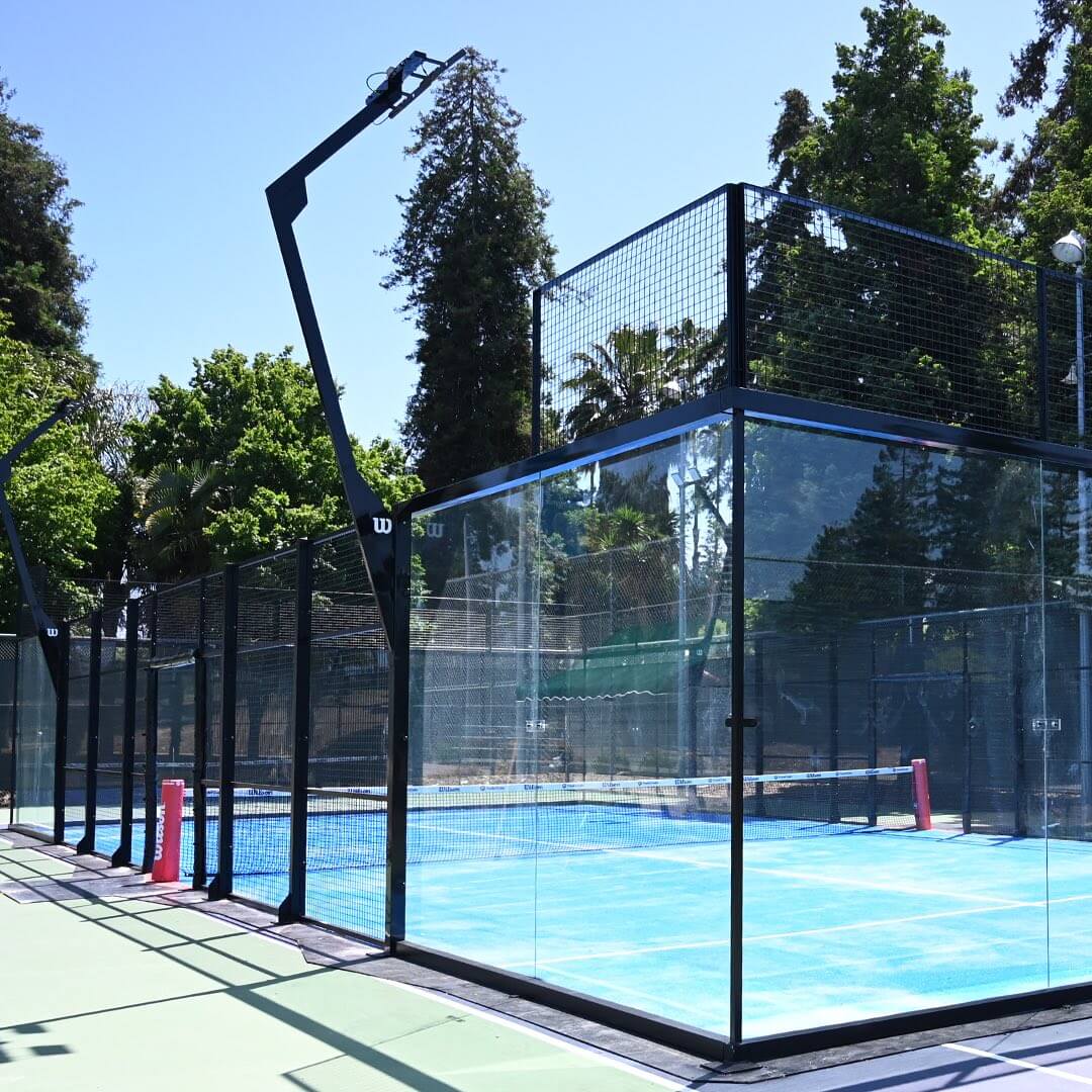 Outdoor glass-enclosed padel court next to tennis courts and tall trees.