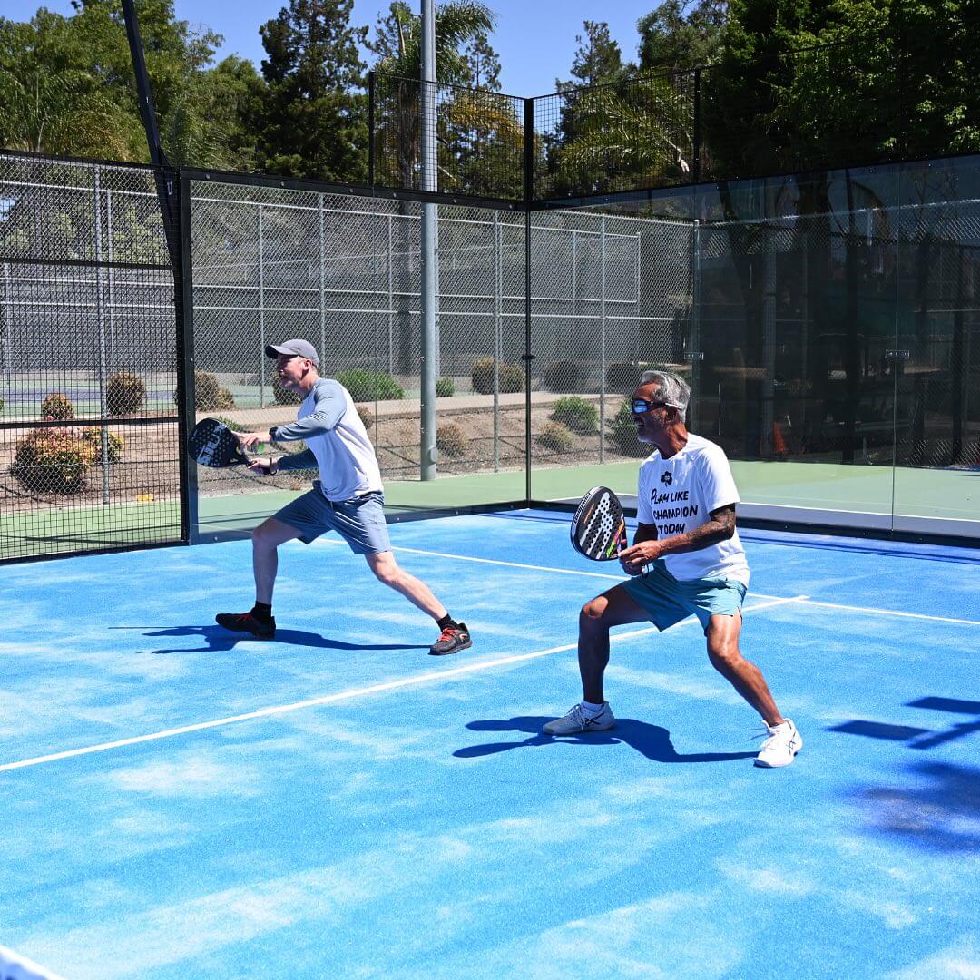 Two players competing on a bright blue padel court outdoors.