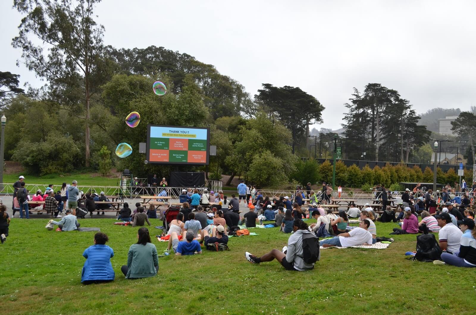Large community event with families seated on grass in front of a big outdoor screen.