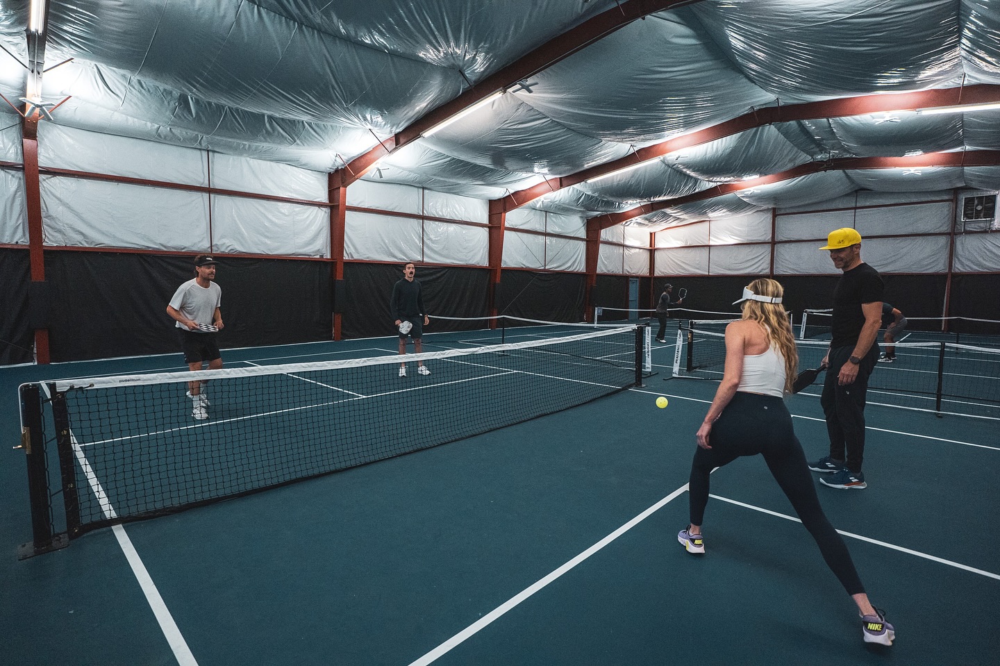 Four players competing on the indoor courts at Bozeman Classic Pickleball Club under bright overhead lighting.