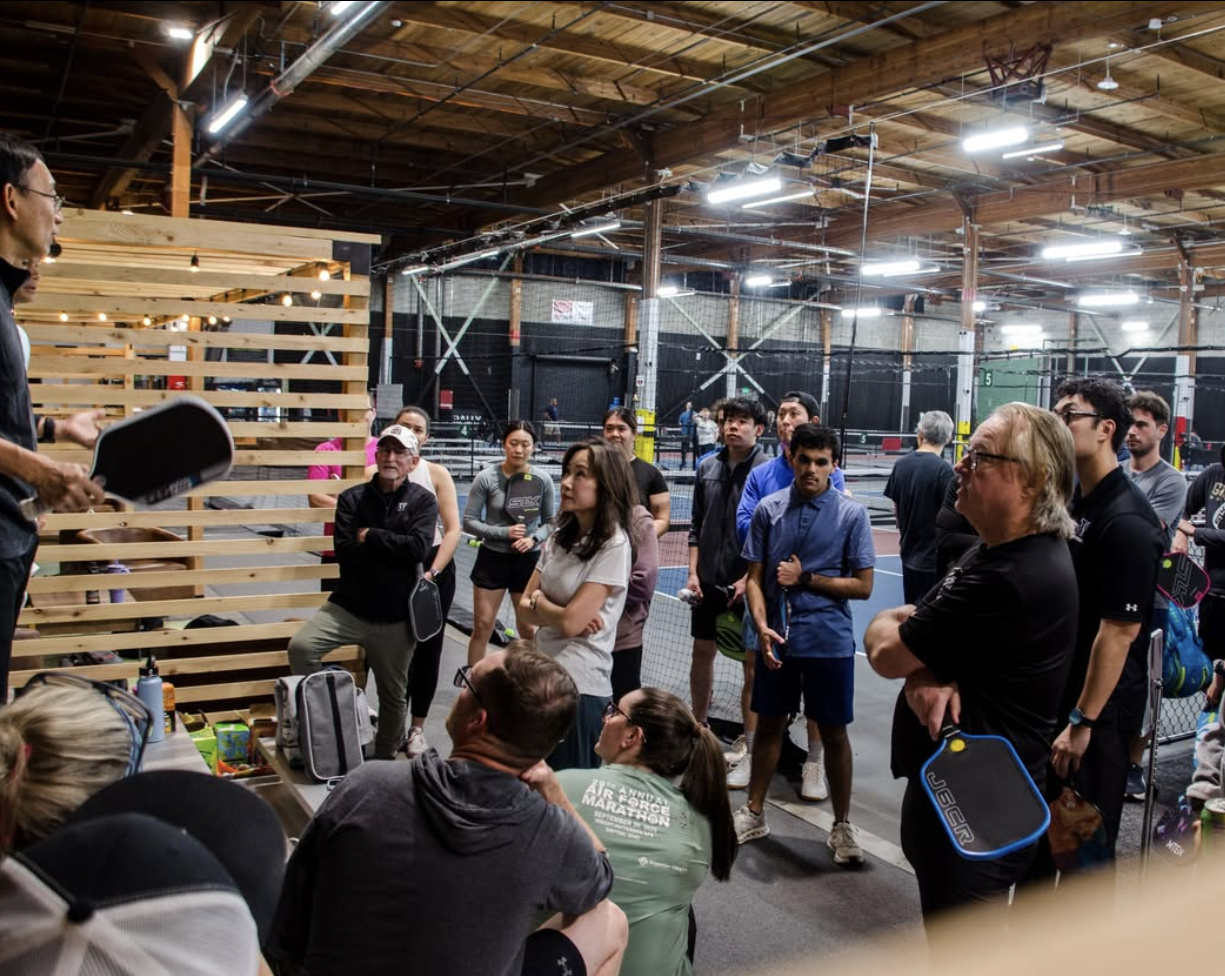 A coach or instructor leading a group clinic or beginner session at Rally Pickleball Club, with players gathered around listening inside the facility.