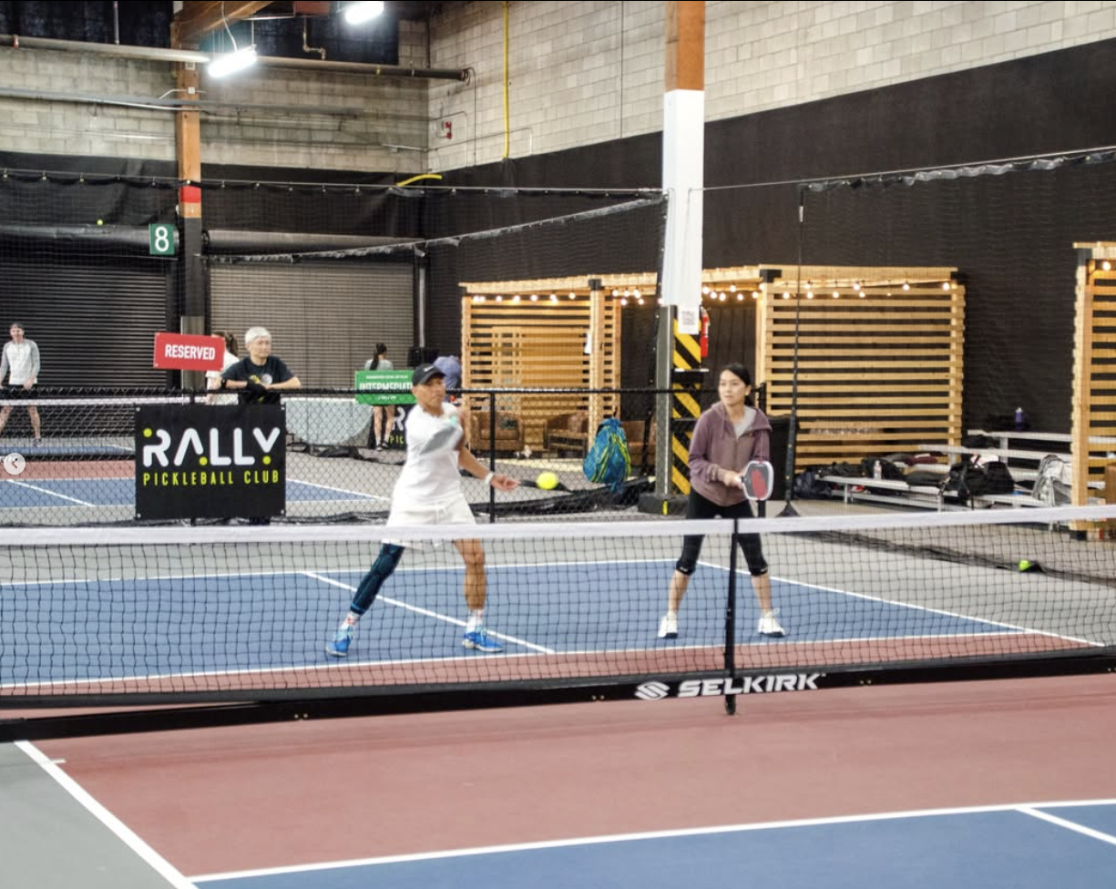 Two players competing on an indoor pickleball court at Rally Pickleball Club, with Selkirk-branded nets and Rally signage visible courtside.