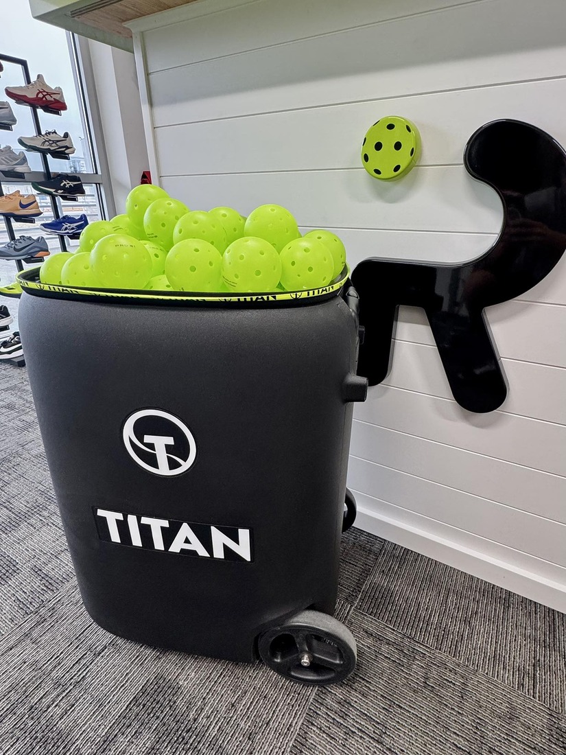 A Titan ball hopper filled with yellow pickleballs in the pro shop at Rally Pickleball Club, with court shoes displayed on a rack in the background.