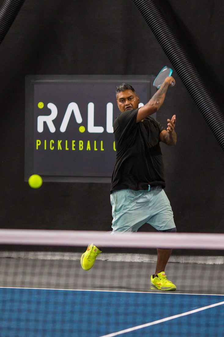 A male pickleball player hitting a backhand shot on an indoor court at Rally Pickleball Club, with the Rally logo visible in the background.