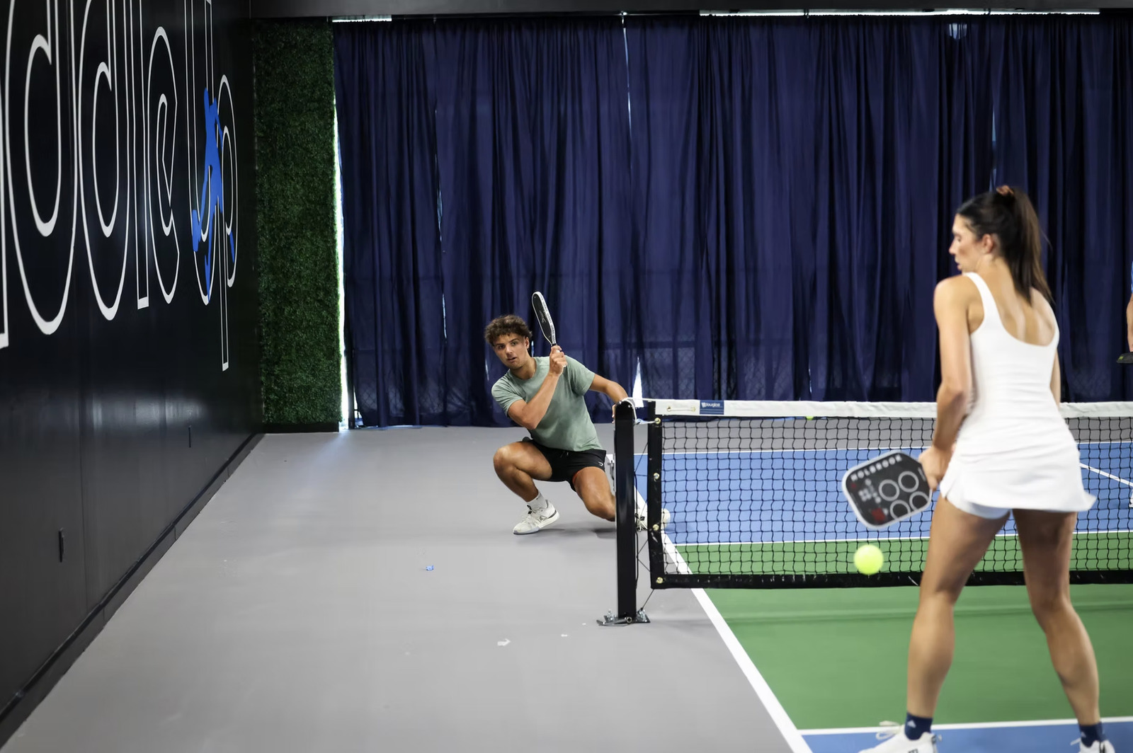 Two players competing in a mixed doubles match on an indoor pickleball court at Paddle Up Pickleball Club, with the Paddle Up logo visible on the back wall.