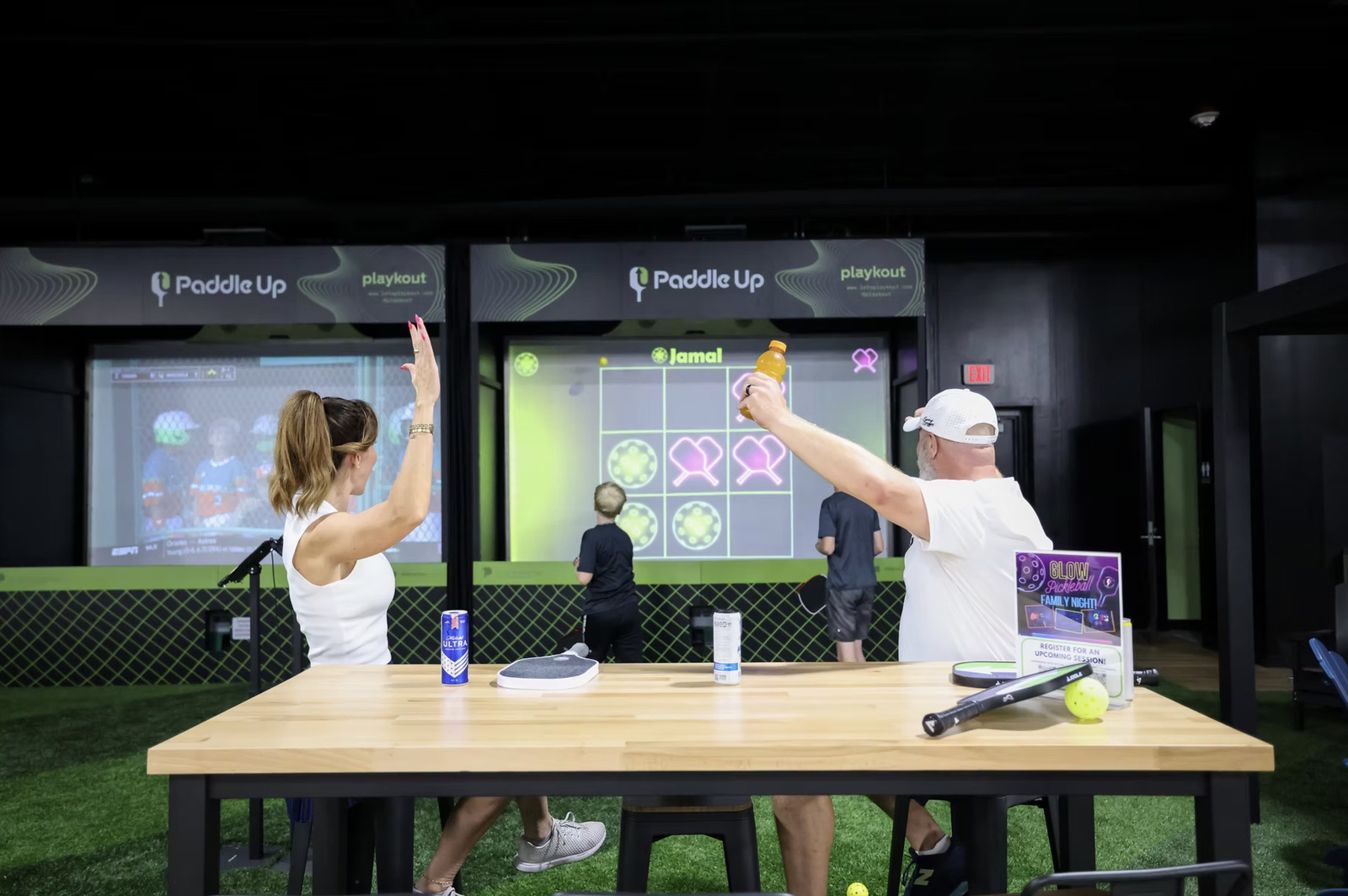Two Paddle Up Pickleball Club members celebrating at a high-top table in front of interactive pickleball simulators during a Glow Pickleball Family Night event.