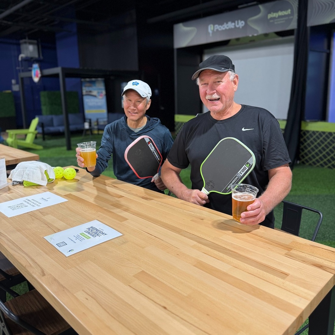 Two Paddle Up Pickleball Club members holding paddles and beers at a high-top table inside the facility, with pickleball simulators and courts visible in the background.