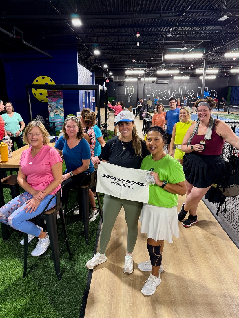 A group of Paddle Up Pickleball Club members gathered inside the facility during an event, with courts visible in the background and a Skechers Pickleball banner in the foreground.