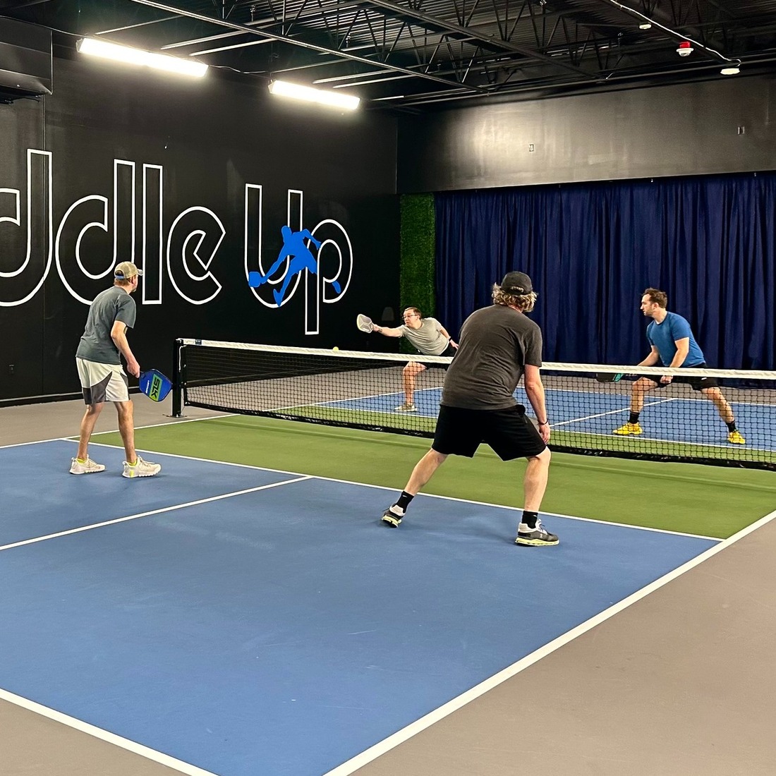 Four players competing in a doubles match on an indoor pickleball court at Paddle Up Pickleball Club, with the Paddle Up logo painted on the back wall.