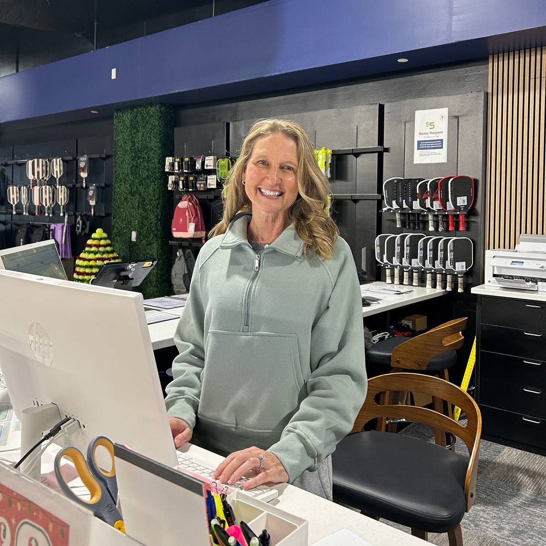 A Paddle Up Pickleball Club staff member smiling at the front desk, with paddles, gear, and apparel displayed in the pro shop behind her.