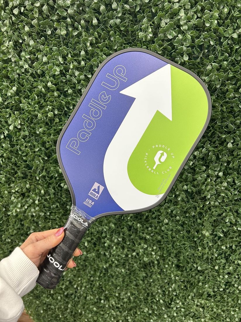 A hand holding a custom JOOLA pickleball paddle branded with the Paddle Up Pickleball Club logo against a green hedge background.