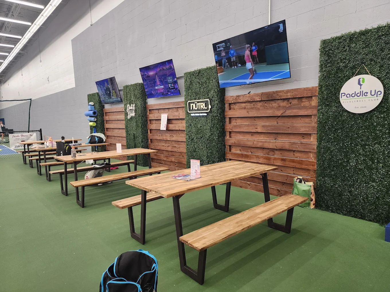 The social lounge area at Paddle Up Pickleball Club featuring wooden picnic tables, hedge wall dividers, and multiple TVs displaying pickleball matches, with the Paddle Up Est. 2023 sign visible.