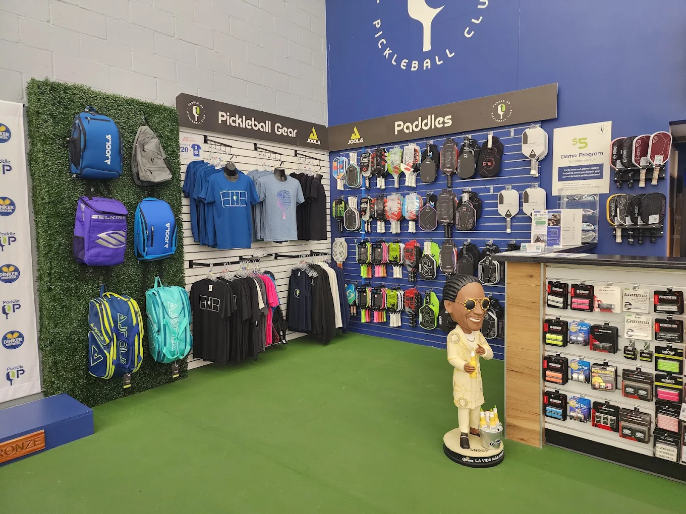 The Paddle Up Pickleball Club pro shop featuring branded apparel, JOOLA paddles, pickleball gear, and a display of Paddle Up Est. 2023 t-shirts.