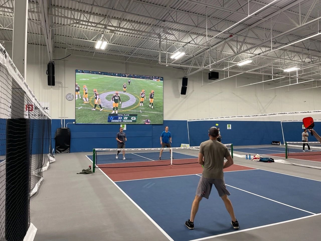 Doubles players rally on a blue and red indoor pickleball court at Pickle Pro Courts, with a football game on a large video wall behind them.