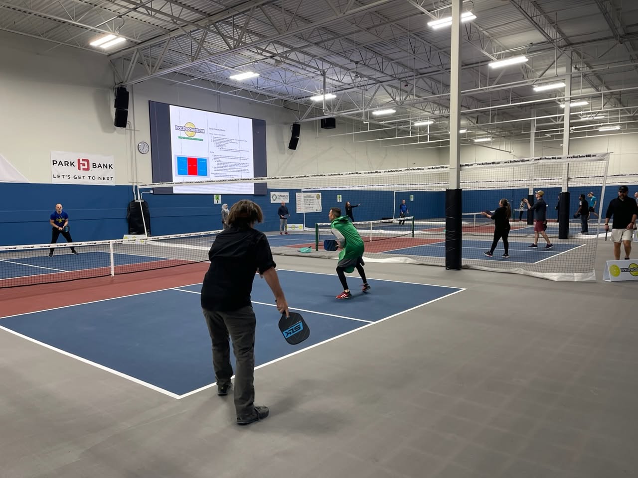 Players fill several indoor courts at Pickle Pro Courts, separated by tall divider nets, as a large screen shows a court diagram.