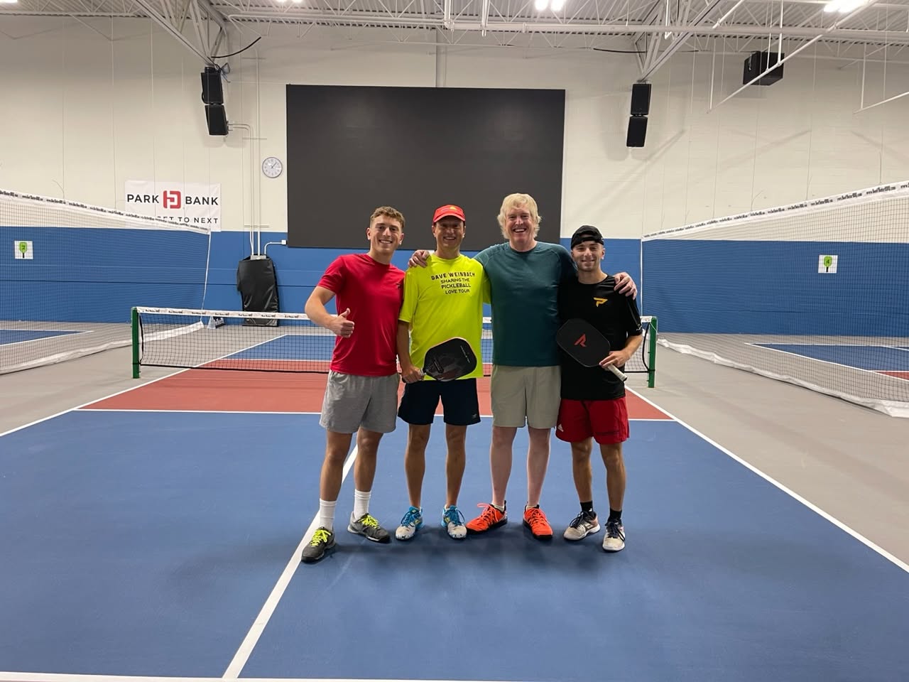 Four smiling players pose arm in arm with paddles on an indoor court at Pickle Pro Courts.