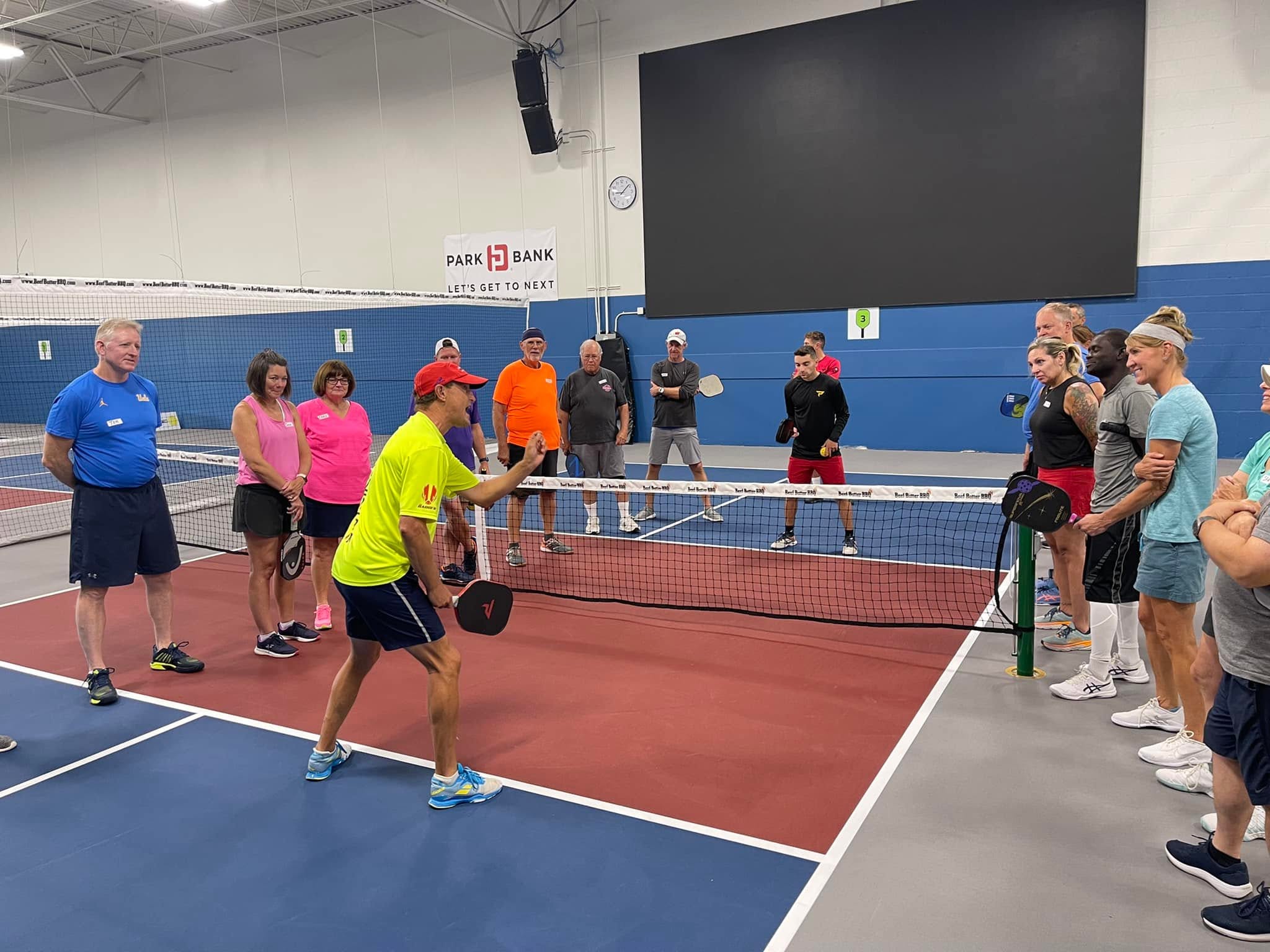 A coach demonstrates a stroke at the net to a group of players gathered around an indoor court at Pickle Pro Courts.