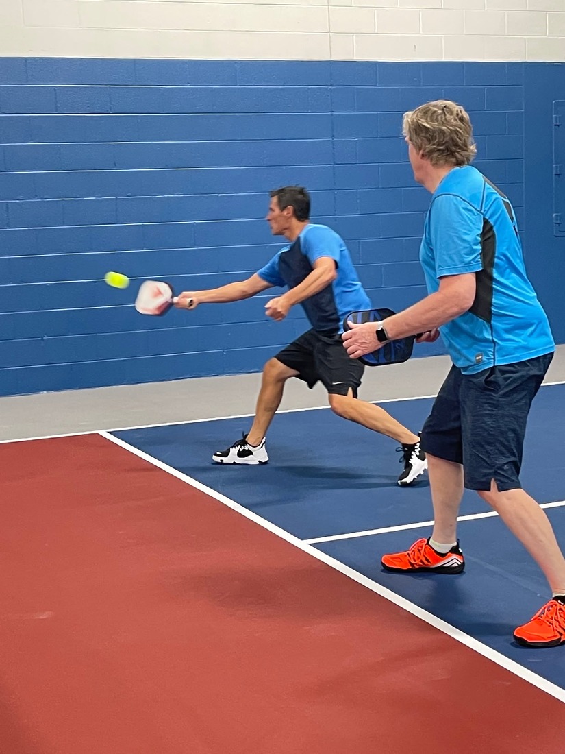 A doubles player reaches for a forehand while his partner watches, on an indoor court at Pickle Pro Courts.
