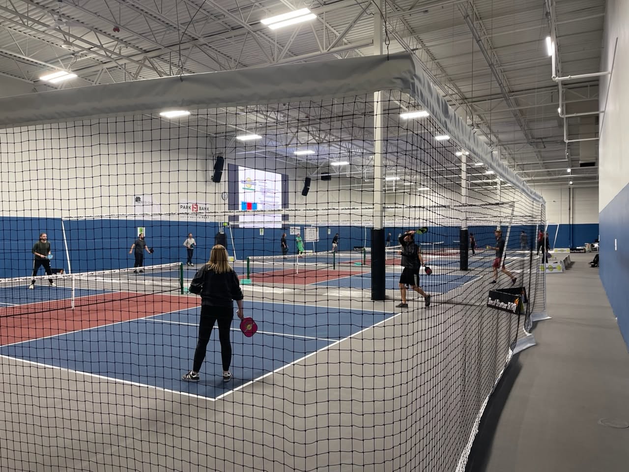 A view through the netting of players on multiple indoor courts at Pickle Pro Courts.