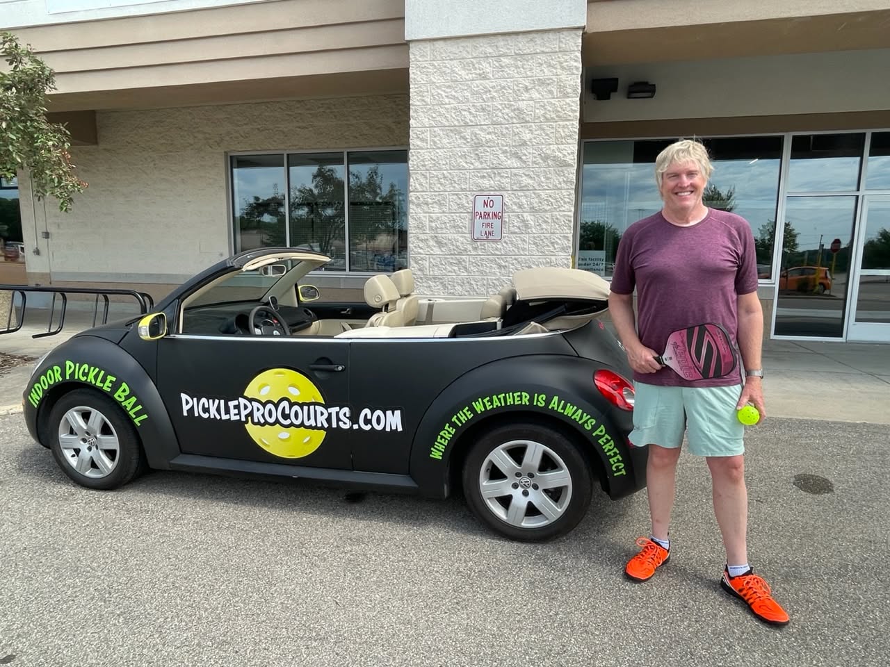 A smiling player holds a paddle and ball beside a black convertible wrapped with PickleProCourts.com branding and the line “Where the weather is always perfect.”